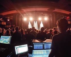 Live sound engineer's perspective from behind two digital mixing consoles at a crowded indoor concert venue. The stage is brightly lit with warm spotlights, illuminating the performing band and the audience dancing in front of the stage.